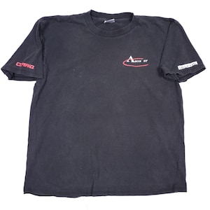 May include: A black short-sleeve t-shirt featuring a red and white graphic on the chest that reads Ascot 07. The right sleeve displays the red text Compaq, while the left sleeve features white text that appears to be partially obscured.