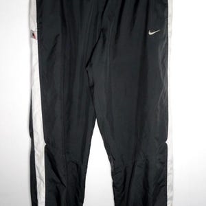 May include: Black athletic trousers featuring a white vertical stripe down each side, an elastic waistband with a drawstring, and a small white Nike swoosh logo on the upper left thigh. The trousers include a small rectangular tag on the right side.