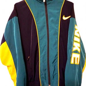 May include: A Nike zip-up track jacket featuring a color-blocked design in dark navy, teal, and bright yellow. The jacket has a high collar, a full-length front zipper, and the word NIKE printed in large yellow letters down the left sleeve.