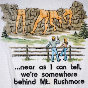 May include: A white t-shirt featuring a graphic illustration of four pairs of human legs emerging from a rocky cliffside. Below, two figures stand by a wooden fence looking at the cliff. The text reads: ...near as I can tell, we're somewhere behind Mt. Rushmore.