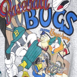 May include: A heather grey t-shirt featuring a graphic print with the text Baseball Bugs Starring Bugs Bunny. The design includes Bugs Bunny wearing a green baseball cap, a baseball, and other cartoon characters in a baseball game setting.