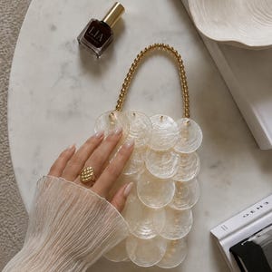 Pode incluir: Uma bolsa feita de discos de concha brancos, circulares e translúcidos sobrepostos, com uma alça de contas douradas. A bolsa está posicionada perto de um pequeno frasco de esmalte escuro e um livro branco com o texto E H O visível.