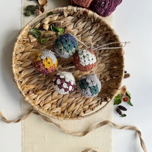 May include: Five crocheted ornaments in a variety of colours and patterns, including green, brown, red, and yellow, are displayed in a woven basket.