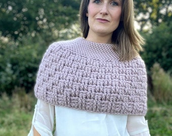 May include: A mauve knitted cowl neck sweater with a textured pattern. The sweater is worn over a white blouse and blue jeans.