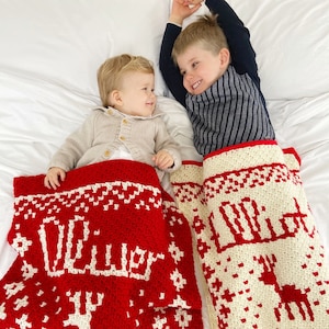 Puede incluir: Dos ni&ntilde;os est&aacute;n tumbados en una cama blanca con sus cabezas en los extremos opuestos de la cama. Cada ni&ntilde;o est&aacute; debajo de una manta de ganchillo con un dise&ntilde;o diferente. La manta de la izquierda es roja con copos de nieve blancos y la palabra "Oliver" en blanco. La manta de la derecha es blanca con copos de nieve rojos y la palabra "Lola" en rojo.