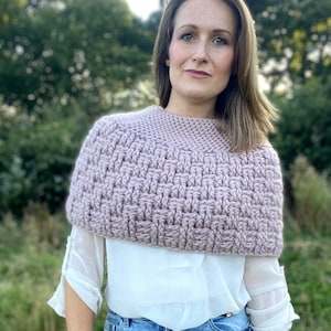 Puede incluir: Un jersey de cuello alto de punto color malva con un patrón texturizado. El jersey se lleva sobre una blusa blanca y unos vaqueros azules.