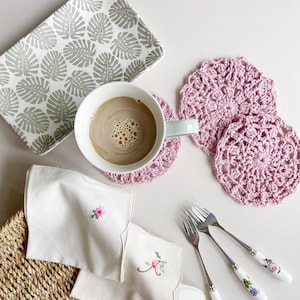 May include: An overhead shot of a light-colored tray with a gray leaf pattern, a mug of coffee, pink crocheted coasters, embroidered napkins, and floral-handled forks. The items are arranged on a light-colored surface.