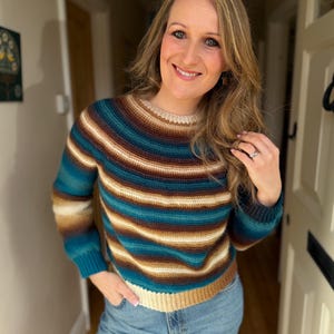 May include: A striped sweater in shades of teal, brown, and cream. The sweater has a crew neck and long sleeves. The person is wearing blue jeans. The sweater's design features horizontal stripes, creating a classic and versatile look.