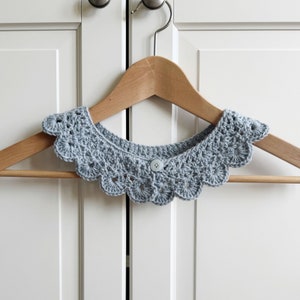 May include: A light blue crocheted collar with a scalloped edge and a single button closure. The collar is hanging on a wooden hanger.