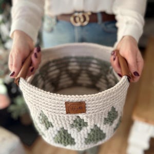 May include: A cream-coloured, crocheted basket with a geometric green pattern. The basket has brown leather handles and a small leather tag. The basket is held by a person.