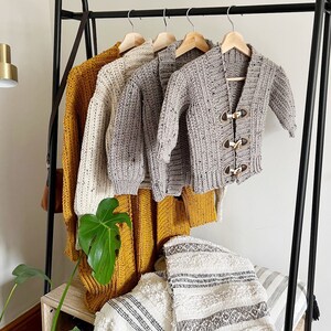 Pu&ograve; includere: Quattro cardigan all'uncinetto in diversi colori appesi a un rack nero. I cardigan sono in tonalit&agrave; di grigio, bianco e giallo. I cardigan sono tutti realizzati con un filato spesso e hanno un design semplice e classico.