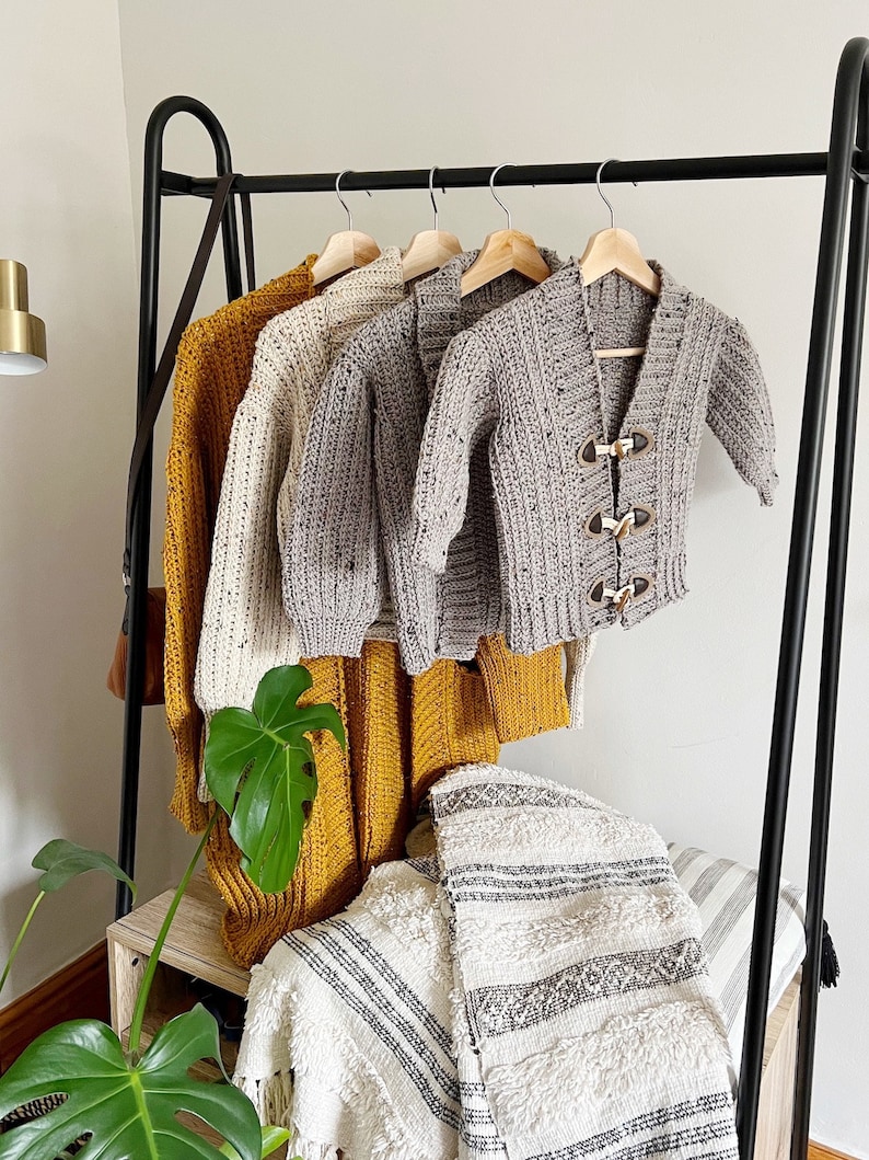 Pu&ograve; includere: Quattro cardigan all'uncinetto in diversi colori appesi a un rack nero. I cardigan sono in tonalit&agrave; di grigio, bianco e giallo. I cardigan sono tutti realizzati con un filato spesso e hanno un design semplice e classico.