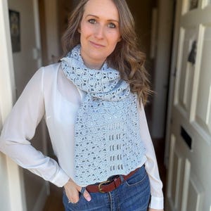 May include: A woman wearing a light blue crocheted scarf with a white shirt and blue jeans.