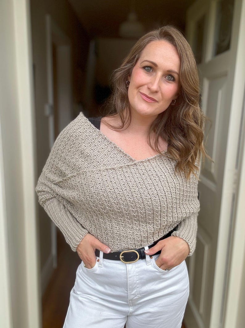 May include: A woman wearing a beige knitted sweater with a V-neck and long sleeves. She is wearing white jeans and a black belt with a gold buckle.