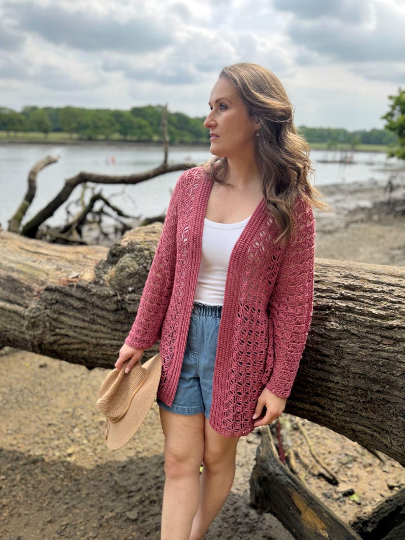 May include: A dusty rose crochet cardigan with an open weave design. The long-sleeved sweater is worn over a white top and denim shorts. A beige sun hat is held in the hand. The background features a river and trees.