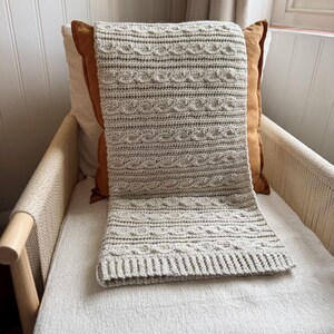 Peut inclure: Une couverture au crochet gris clair avec un motif en tricot torsad&eacute;, drap&eacute;e sur une chaise. La couverture a un aspect textur&eacute; et est faite d'un fil doux. La chaise a un cadre en bois clair et une assise tiss&eacute;e.