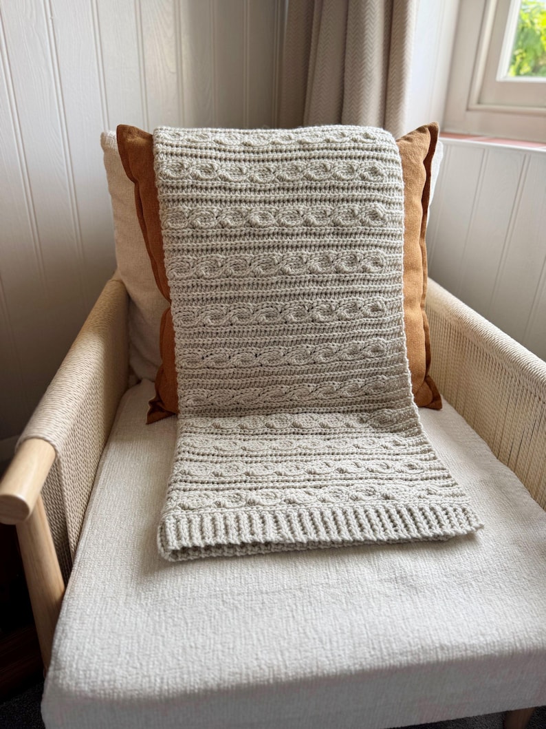 Peut inclure: Une couverture au crochet gris clair avec un motif en tricot torsad&eacute;, drap&eacute;e sur une chaise. La couverture a un aspect textur&eacute; et est faite d'un fil doux. La chaise a un cadre en bois clair et une assise tiss&eacute;e.