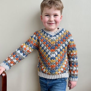 May include: A child wearing a colorful crocheted sweater with a geometric pattern in shades of blue, orange, and yellow. The sweater has a V-neck and long sleeves.