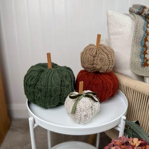 May include: Four knitted pumpkins in shades of green, brown, orange, and cream, each with a cinnamon stick stem. The cream pumpkin has a green velvet ribbon. The pumpkins are displayed on a white, two-tiered side table, creating a cozy autumn display.