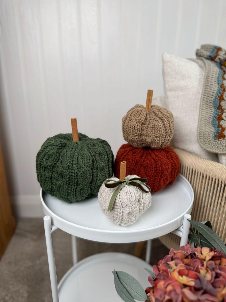 May include: Four knitted pumpkins in shades of green, brown, orange, and cream, each with a cinnamon stick stem. The cream pumpkin has a green velvet ribbon. The pumpkins are displayed on a white, two-tiered side table, creating a cozy autumn display.