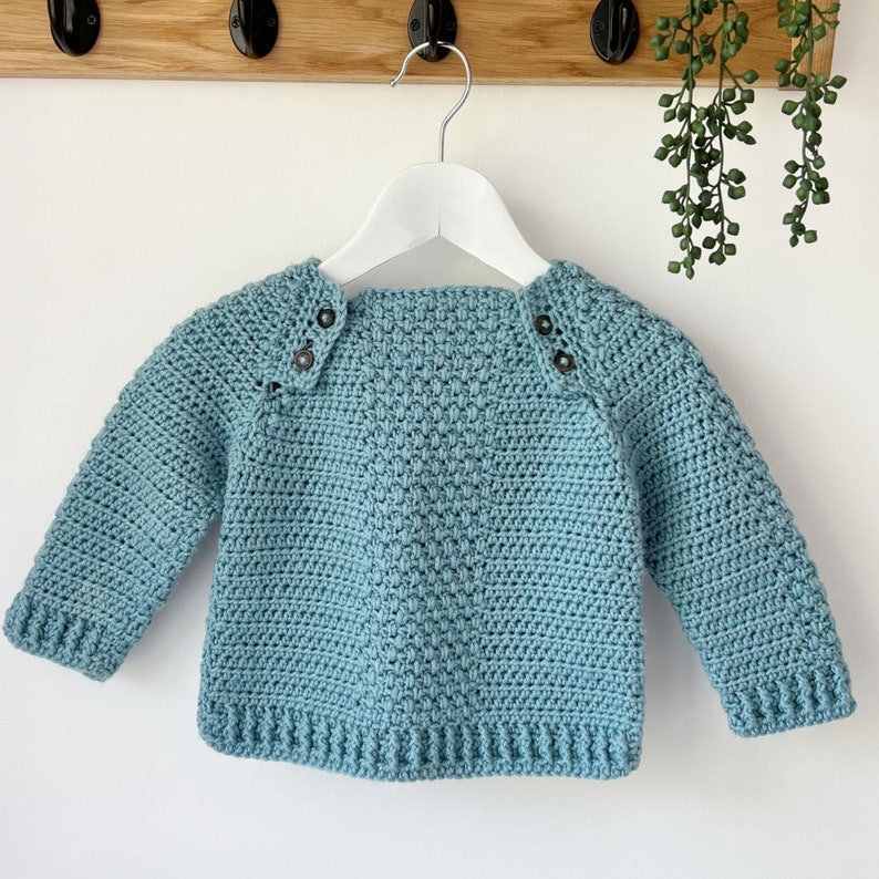 May include: A light blue crocheted sweater with black buttons on the shoulders. The sweater is hanging on a white hanger against a white wall.