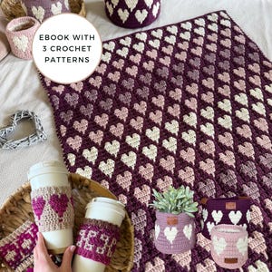 May include: A collection of handmade crochet items in shades of purple, pink, and white. Includes a blanket, baskets, coffee cup sleeves, and a small plant pot, all featuring heart patterns. The text "EBOOK WITH 3 CROCHET PATTERNS" is visible.