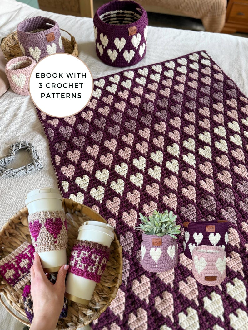 Op de afbeelding: Een verzameling handgemaakte haakartikelen in tinten paars, roze en wit. Inclusief een deken, manden, koffiekokers en een kleine plantenpot, allemaal met hartpatronen. De tekst "EBOOK WITH 3 CROCHET PATTERNS" is zichtbaar.