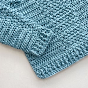 May include: A close-up of a blue crocheted fabric with a textured pattern. The fabric is folded over on itself, creating a seam.