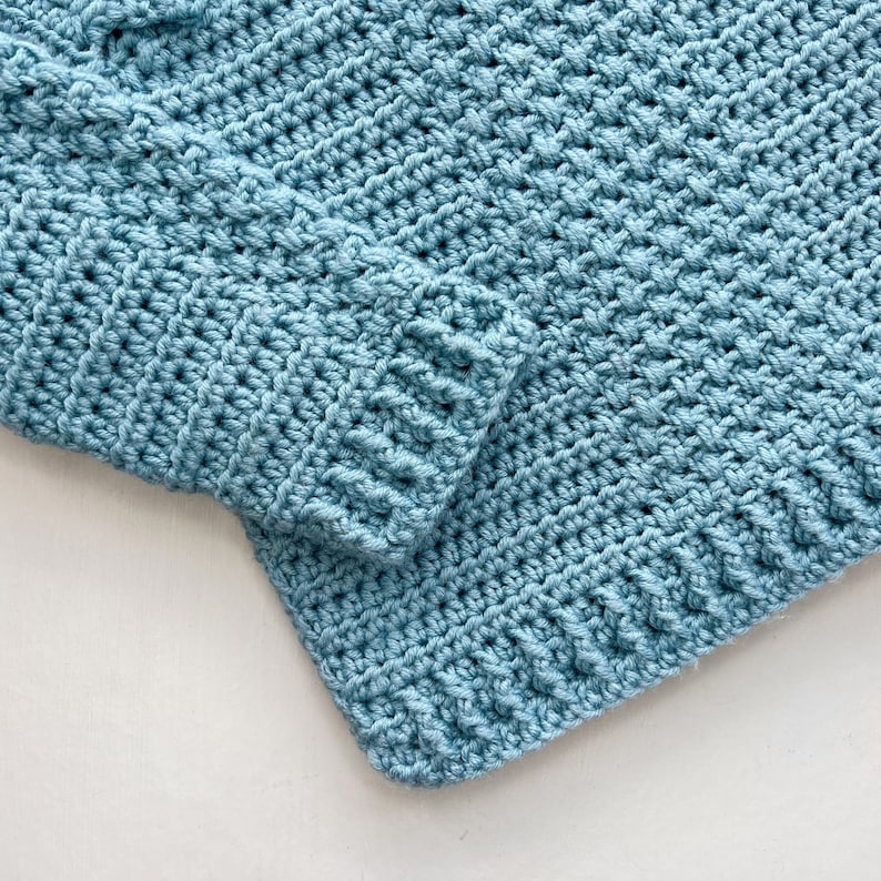 May include: A close-up of a blue crocheted fabric with a textured pattern. The fabric is folded over on itself, creating a seam.