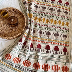 May include: A cream-coloured crochet blanket featuring horizontal rows of autumn-themed motifs including orange pumpkins, red mushrooms, brown acorns, and yellow leaves, finished with a sage green border and intricate stitch patterns throughout.