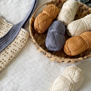 Puede incluir: Un bolso tote de ganchillo con cuerpo color crema y asas grises, acompa&ntilde;ado de una cesta tejida que contiene seis ovillos de lana en tonos crema, amarillo mostaza y azul pizarra, con un ovillo de lana crema adicional colocado fuera.