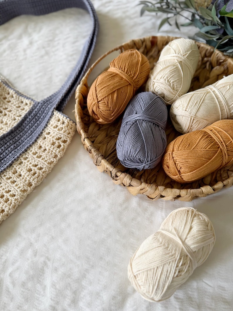 Puede incluir: Un bolso tote de ganchillo con cuerpo color crema y asas grises, acompa&ntilde;ado de una cesta tejida que contiene seis ovillos de lana en tonos crema, amarillo mostaza y azul pizarra, con un ovillo de lana crema adicional colocado fuera.