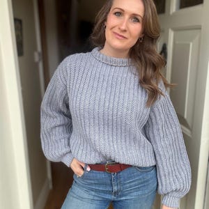 May include: A woman wearing a light gray knitted sweater with a brown leather belt and blue jeans.