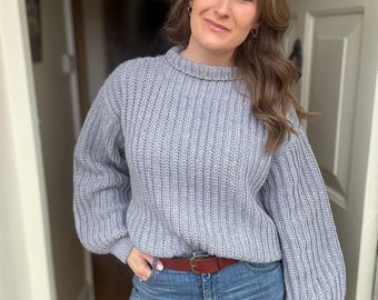 Könnte beinhalten: Ein hellgrauer Rippstrickpullover mit Rundhalsausschnitt und langen Ärmeln. Der Pullover wird mit einer blauen Jeans und einem braunen Ledergürtel mit goldfarbener Schnalle kombiniert. Die Person steht in einem Türrahmen.