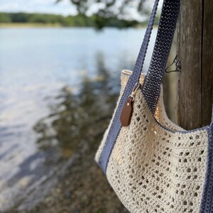 Puede incluir: Un bolso de ganchillo con un patr&oacute;n calado en color blanco roto, correas y bordes en gris s&oacute;lido. Una peque&ntilde;a etiqueta de cuero sint&eacute;tico marr&oacute;n est&aacute; sujeta a la correa con un broche de metal dorado. El bolso tiene un dise&ntilde;o estructurado.