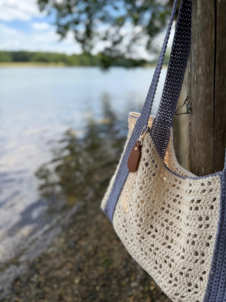 Puede incluir: Un bolso de ganchillo con un patr&oacute;n calado en color blanco roto, correas y bordes en gris s&oacute;lido. Una peque&ntilde;a etiqueta de cuero sint&eacute;tico marr&oacute;n est&aacute; sujeta a la correa con un broche de metal dorado. El bolso tiene un dise&ntilde;o estructurado.