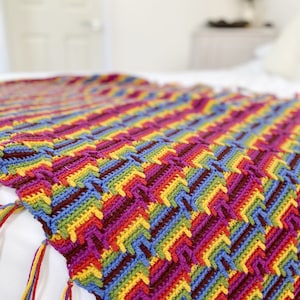 Puede incluir: Una colorida manta de ganchillo hecha a mano con un patrón de chevrón. La manta presenta una vibrante gama de colores que incluyen rojo, amarillo, azul y morado. Los bordes están acabados con borlas en colores a juego.