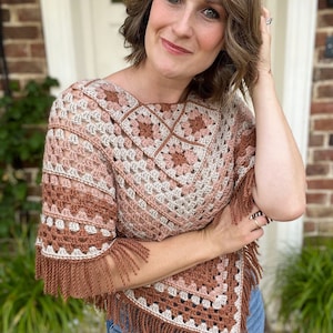 Puede incluir: Un poncho de crochet con un patr&oacute;n geom&eacute;trico en tonos marrones, rosas y beige. El poncho tiene flecos en los bordes y lo lleva una persona con el pelo casta&ntilde;o.