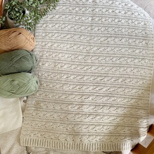 Peut inclure: Une couverture tricot&eacute;e beige clair avec un motif en torsades. La couverture est rectangulaire et poss&egrave;de une bordure c&ocirc;tel&eacute;e. Plusieurs pelotes de laine beiges, vertes et marron sont &agrave; c&ocirc;t&eacute; de la couverture, avec une petite plante en pot.