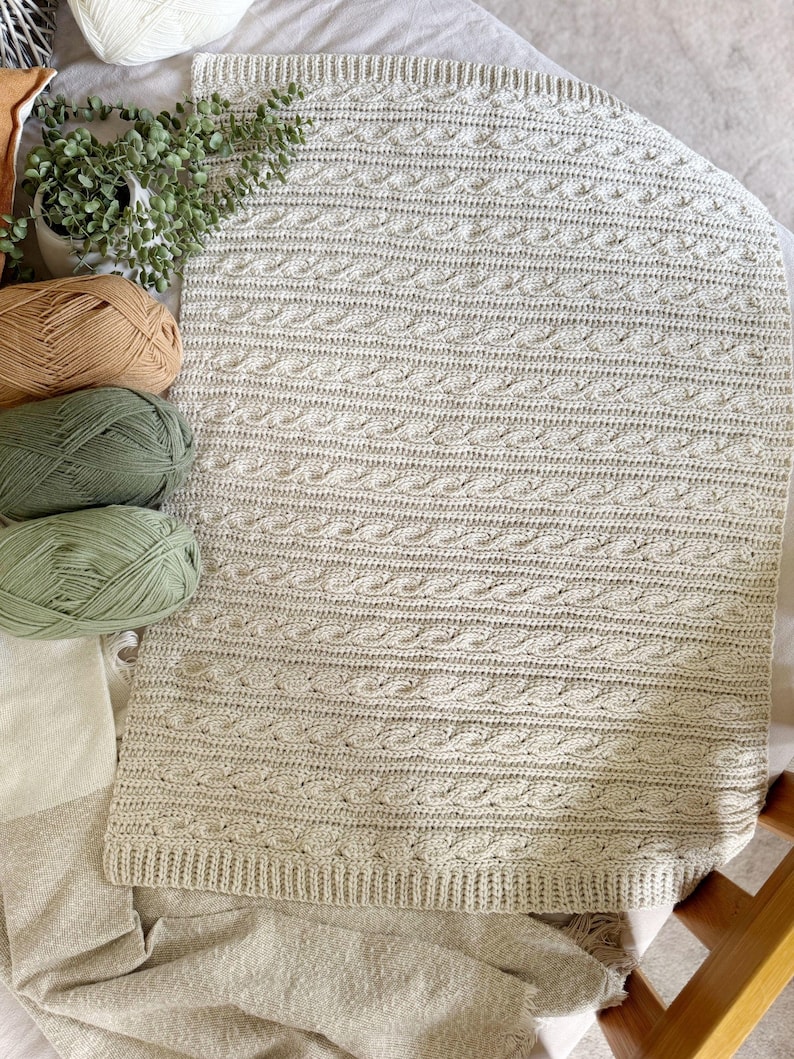Peut inclure: Une couverture tricot&eacute;e beige clair avec un motif en torsades. La couverture est rectangulaire et poss&egrave;de une bordure c&ocirc;tel&eacute;e. Plusieurs pelotes de laine beiges, vertes et marron sont &agrave; c&ocirc;t&eacute; de la couverture, avec une petite plante en pot.