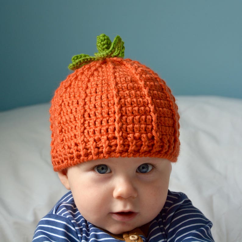 May include: A baby wearing a crocheted orange pumpkin hat with green leaves. The hat is made with a textured stitch and covers the baby's head and ears.