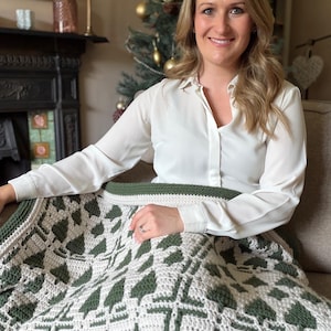 May include: A crochet blanket featuring a repeating pattern of dark green pine trees on a light cream background. The blanket has a solid dark green border followed by a light cream edge, showcasing a textured stitch design throughout the fabric.