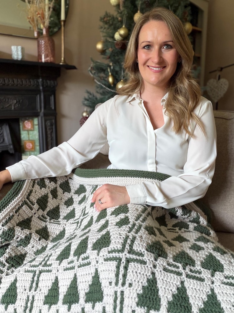 May include: A crochet blanket featuring a repeating pattern of dark green pine trees on a light cream background. The blanket has a solid dark green border followed by a light cream edge, showcasing a textured stitch design throughout the fabric.