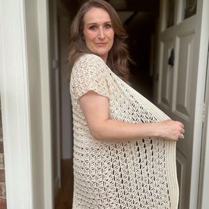 May include: Cream-colored, open-weave crochet cardigan with short sleeves. The cardigan features a geometric pattern and a ribbed border. The garment is lightweight and appears to be handmade. The style is a relaxed fit, suitable for layering.