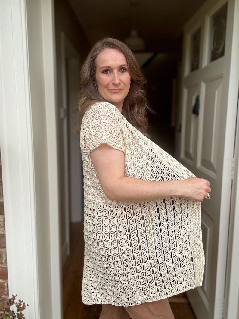 May include: Cream-colored, open-weave crochet cardigan with short sleeves. The cardigan features a geometric pattern and a ribbed border. The garment is lightweight and appears to be handmade. The style is a relaxed fit, suitable for layering.