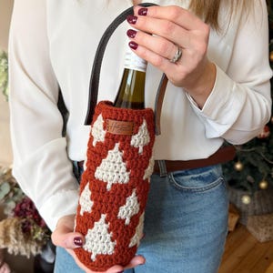 May include: A crocheted wine bottle holder in a rust-colored hue, adorned with white tree motifs. It features a black leather handle and a small brown tag. The holder is designed to carry a standard-sized wine bottle.