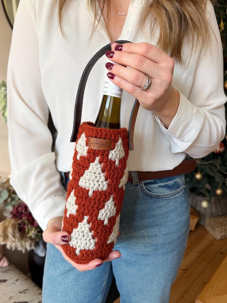 May include: A crocheted wine bottle holder in a rust-colored hue, adorned with white tree motifs. It features a black leather handle and a small brown tag. The holder is designed to carry a standard-sized wine bottle.