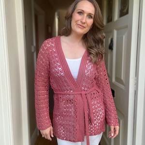 May include: A dusty rose crochet cardigan with long sleeves and a matching belt. The open-weave design reveals a white top underneath. The cardigan is worn with white pants.