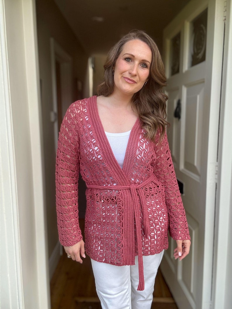 May include: A dusty rose crochet cardigan with long sleeves and a matching belt. The open-weave design reveals a white top underneath. The cardigan is worn with white pants.