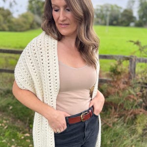May include: Cream-colored, open-weave crochet shawl. The shawl is draped over the shoulders. The model is wearing a tan tank top, dark jeans, and a brown leather belt with a gold buckle. The background is a green field and a wooden fence.